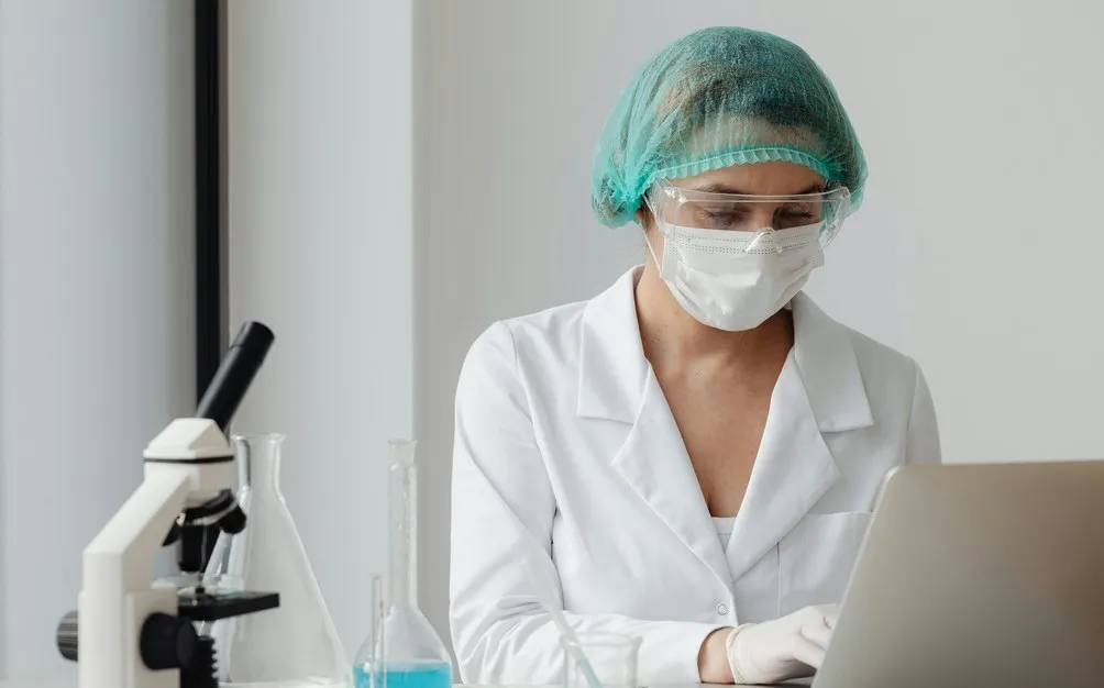 A lab researcher in a hairnet and mask working at a laptop beside a microscope